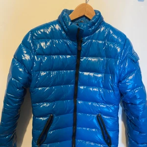 Blå Moncler pufferjacka (S) - Snygg blå pufferjacka från Moncler i storlek S med glansig finish och svarta dragkedjor. Jackan har hög krage, två fickor framtill och en ärmficka med Moncler-logga. Insidan har ett unikt serietryck. Perfekt för dig som vill sticka ut i vinterkylan.