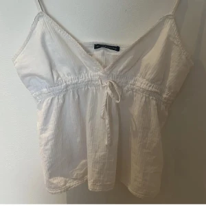 Vit linnetopp med knyt från Brandy Melville - Söt vit linnetopp från Brandy Melville med smala axelband och v-ringning. Toppen har en knytning framtill och resår under bysten för en snygg passform. Tillverkad i mjuk bomull, perfekt för varma dagar.