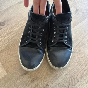 Svarta sneakers från Lanvin - Snygga svarta sneakers från Lanvin med ovandel i mocka och detaljer i skinn. Skorna har rund tå, snörning med metallöljetter och vit platt sula. Perfekta för dig som gillar stilrena och klassiska sneakers med exklusiv känsla.