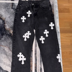 Chroma heart / Levi's 550 svarta jeans - Svarta Levi's 550 jeans med vita korsapplikationer över hela byxorna. Jeansen har klassisk femficksdesign, silverfärgade nitar och knapp med unik detalj. Råa, fransiga benslut för en edgy look. Materialet är robust denim.