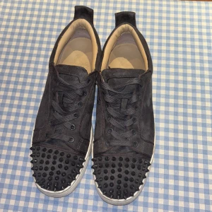 Svarta mockasneakers med nitar Louboutin - Svarta sneakers i mocka från Christian Louboutin med coola svarta nitar på tån och klassisk röd sula. Skorna har snörning och vit platt sula. Insidan är beige och de har en modern, edgy vibe som sticker ut. Ganska tajta då det egentligen är kostymskor. Men de funkar för mig som har ungefär 43,5 och är perfekta i vardagslivet!