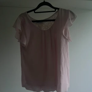 Ljusrosa blus med spets från Creme Fraiche - Supersöt ljusrosa blus från Creme Fraiche med volangärmar och v-ringning. Baksidan har en snygg öppen detalj med spetsinsats som ger en extra touch. Luftigt material och flowig passform, perfekt för dig som gillar romantisk stil.