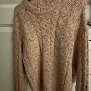 Beige stickad kabeltröja - Säljer en mysig rosa stickad tröja med grovt kabelstickat mönster och rund hals. Perfekt för kyliga dagar och enkel att styla med jeans eller kjol. Tröjan har långa ärmar och en avslappnad passform.
