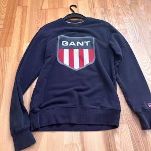 Marinblå sweatshirt från GANT S - Snygg marinblå sweatshirt från GANT i storlek S. Tröjan har ett stort tryckt GANT-emblem med röda och vita ränder på bröstet och ett mindre broderat märke på ärmen. Rund hals och ribbade muddar. Perfekt för dig som gillar klassisk och sportig stil.