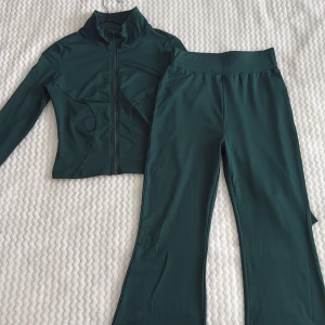 Mörkgrönt träningsset med zip - Snyggt mörkgrönt träningsset med långärmad zip-jacka och matchande byxor med utsvängda ben. Jackan har hög krage och dragkedja framtill. Materialet är stretchigt, tröjan är använd några gånger men aldrig byxorna. 