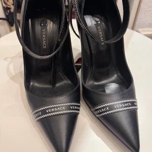 Svarta Versace pumps med logoband - Snygga svarta pumps från Versace i skinn med spetsig tå och hög klack. Skorna har ett svartvitt band med Versace-logga över foten och runt vristen, samt guldfärgad Medusa-detalj på sulan. Perfekta för dig som vill sticka ut med lyxig designerstil.