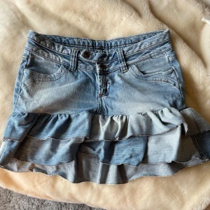 Jeanskjol med volanger och fickor - Snygg ljusblå jeanskjol med dubbla volanger nertill och klassiska jeansfickor både fram och bak. Kjolen har bälteshällor, dubbla knappar och dragkedja framtill. Perfekt för dig som gillar en cool och lekfull stil. Köpt här på plick men passar inte mig