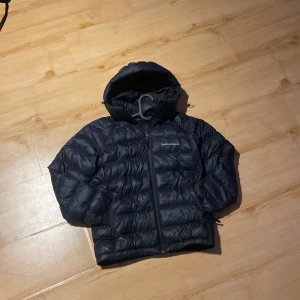 Svart dunjacka Peak Performance 150 - Svart dunjacka från Peak Performance med huva och broderad logga på bröstet. Jackan har quiltad design, dragkedja framtill och är perfekt för kalla dagar. Materialet är syntet och dun, och passformen är normal. Snygg och funktionell för dig som gillar stil och värme.