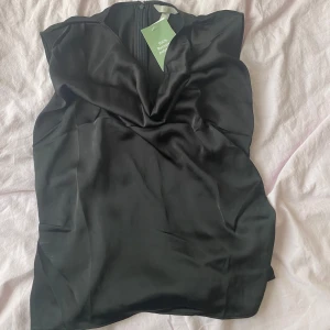 Svart ärmlös blus i satin - Snygg svart blus i glansigt satinmaterial. Blusen har en draperad v-ringad front och är ärmlös för en clean look. Perfekt att styla med jeans eller kjol. Stängs med dragkedja bak. Aldrig använd prislapp kvar. 