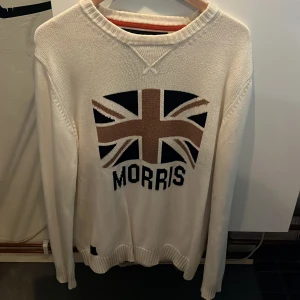 Vit stickad tröja från Morris - Vit stickad tröja från Morris i storlek Large med Union Jack-flagga och Morris-tryck framtill. Tröjan har rund halsringning, ribbade muddar och lång ärm. Perfekt för dig som gillar klassisk brittisk stil och vill ha något snyggt och bekvämt.