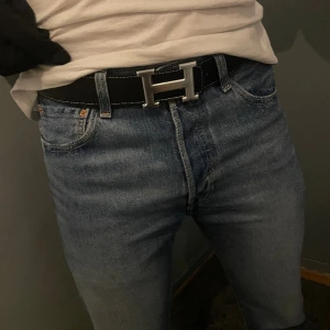 Svart bälte med H-spänne - Säljer ett svart bälte med ikoniskt H-spänne i silverfärgad metall. Bältet har vita kontrastsömmar längs kanterna. Perfekt accessoar för att lyfta din outfit med en lyxig touch.