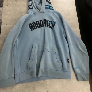 Blå hoodie från Hoodrich med tryck - Säljer en ljusblå hoodie från Hoodrich med svart och blått tryck framtill och text på ryggen. Tröjan har stor huva, känguruficka och broderade detaljer på ärmen. Perfekt för dig som gillar streetwear och vill sticka ut lite extra.