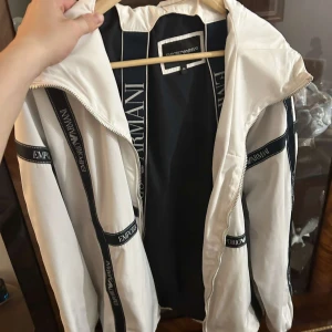 Vit vindjacka från Emporio Armani - Snygg vit vindjacka från Emporio Armani med svarta detaljer och logoband längs ärmar och rygg. Jackan har huva, hel dragkedja och sportig look. Materialet är lätt och känns fräscht, perfekt för dig som gillar stilrena streetwear-vibbar. Enda deffenten är Små hål på vänster arm som knappt syns, går säkert att laga. Den är använd cirka 5 gånger.  Ny pris 530euro. Peis går att diskutera
