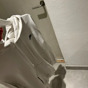 Vit hoodie från Nike med huva - Vit hoodie från Nike med klassisk svart logga på bröstet. Tröjan har huva med snörning och en stor magficka framtill. Mjuk insida och relaxed passform, perfekt för chill dagar. Snygg och enkel design som funkar till det mesta.