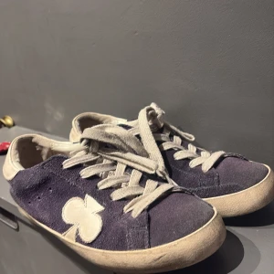 Mörkblå sneakers med klöverdetalj - Snygga mörkblå sneakers med vit klöverdetalj på sidan och vita snören. Skorna har en klassisk rund tå och platt sula. Ovandelen är i mocka och detaljerna i vitt ger en cool kontrast. Perfekta för dig som gillar unika detaljer på dina skor.