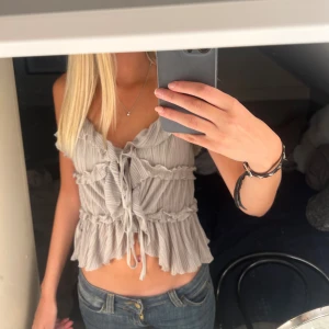  volangig blus med snörning - Supersöt blus med volanger och knytning framtill. Passar till både jeans och kjol för en trendig och fin outfit! Storlek S från New Yorker Amisu. Pris kan diskuteras och hör av er om ni har frågor. Toppen är oanvänd. Den är begie grå 