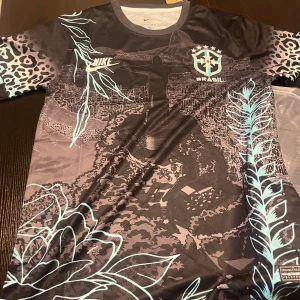 Nike Brasil fotbollströja svart/blå S - Snygg Brasilien fotbollströja från Nike med svart bas och ljusblå detaljer. Tröjan har korta ärmar, leopardmönster på axlarna och grafiskt tryck med Kristusstatyn och växtmotiv. Tillverkad i Dri-FIT material som andas.