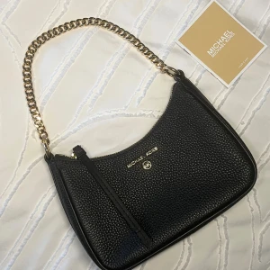 Svart axelväska från Michael Kors - Stilren svart axelväska från Michael Kors i präglat skinn med guldfärgad kedja som axelrem. Väskan har dragkedja upptill och diskret logga i guld på framsidan. Perfekt storlek för det viktigaste och ger en lyxig känsla till din outfit.