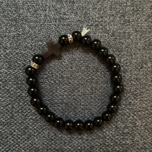 Svart pärlarmband med kors - Stilrent svart armband med glansiga pärlor och ett svart kors som detalj. Armbandet har även små silvriga detaljer mellan pärlorna. Perfekt accessoar för dig som gillar minimalistisk och cool stil.