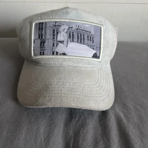 Vit keps från Emilio Pendenza - Säljer en vit keps från Emilio Pendenza med mesh på baksidan och en unik patch framtill med svartvitt motiv. Kepsen har böjd skärm och är tillverkad i ett mjukt material som känns skönt på huvudet. Perfekt för dig som vill sticka ut med en cool accessoar.