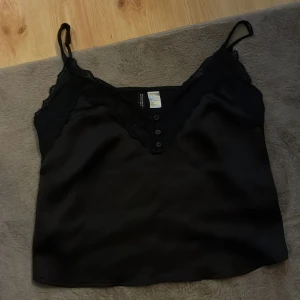 Svart linnetopp med spets från Divided - Säljer en svart linnetopp från Divided med smala axelband och v-ringning dekorerad med spets. Toppen har knappar framtill och är gjord i ett glansigt, satinliknande material som känns riktigt mjukt mot huden. Perfekt för att lyfta vilken outfit som helst.