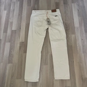 Vita jeans från Nudie Jeans - Snygga vita jeans från Nudie Jeans med klassisk femficksdesign och diskret logga på bakfickan. Byxorna har raka ben och är tillverkade i ett slitstarkt bomullstyg. Perfekta för dig som gillar en clean och stilren look.