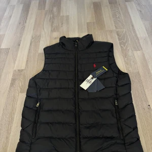 Svart dunväst från Polo Ralph Lauren - Svart quiltad dunväst från Polo Ralph Lauren med röd broderad logga på bröstet. Västen har hög krage, två dragkedjefickor och hel dragkedja framtill. Perfekt för lager-på-lager och snygg till både hoodie och skjorta.