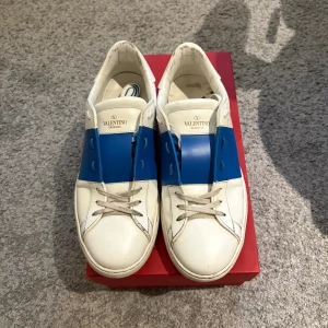 Valentino Garavani Open sneakers blå/vit - Snygga Valentino Garavani Open sneakers i vitt skinn med en bred blå läderdetalj över mitten. Klassisk rund tå, vita snören och diskret logga på plösen. Sulan är platt och har en cool nitar-detalj baktill. Perfekta för dig som gillar exklusiva och trendiga sneakers.