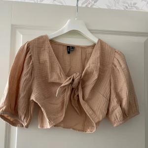 Beige croppad blus med knyt - Superfin beige croppad blus med puffiga trekvartsärmar och knyt framtill. Tunt, luftigt bomullstyg med lätt struktur och v-ringad hals. Perfekt för en chill och trendig look.