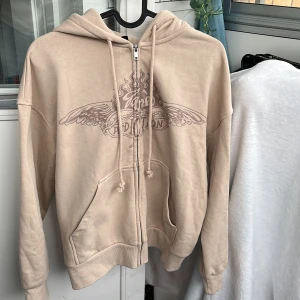 Beige hoodie med tryck och dragkedja  - Beige hoodie från HM med text på bröstet. Tröjan har huva med snörning, dragkedja framtill och stora fickor. Mjuk och skön, perfekt för chill dagar. Passar dig som gillar avslappnad stil.