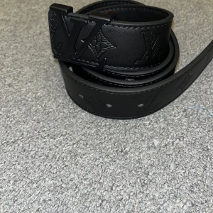 Svart bälte - Snyggt svart skärp  i skinn med präglade loggor och monogramdetaljer längs hela bältet. Mattsvart spänne . Perfekt accessoar för att lyfta din outfit med en lyxig touch.