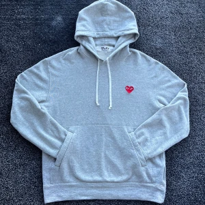 CDG Hoodie - Grå hoodie från Comme des Garçons Play i storlek XL. Klassisk modell med huva, snörning och känguruficka. På bröstet sitter det ikoniska röda hjärtat med ögon. Mjuk bomullskvalitet och långärmad passform. Skick 8/10, använd, storlek XL men passar mer som L, äkta. Pris går att diskutera✅