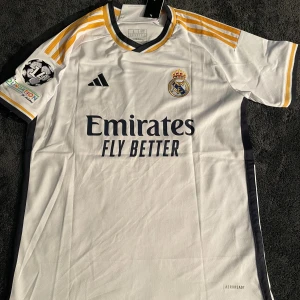 Real Madrid Valverde #5 fotbollströja - Snygg Real Madrid matchtröja med Valverde och nummer 5 på ryggen. Vit bas med mörkblå detaljer och guldiga ränder på axlarna. Broderat klubbmärke, Champions League-märke på ärmen och 'Emirates Fly Better' tryck på bröstet. Tillverkad i lätt polyester med Aeroready-teknologi.