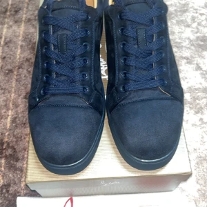 Mörkblå sneakers från Christian Louboutin - Snygga mörkblå sneakers från Christian Louboutin i mocka med klassisk röd sula och broderad logga på hälen. Skorna har rund tå, platt sula och snörning. Perfekta för dig som vill sticka ut med exklusiva detaljer och lyxig känsla.