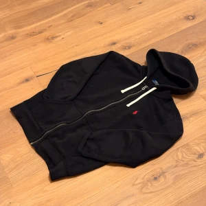 Svart hoodie från Polo Ralph Lauren - Svart hoodie från Polo Ralph Lauren med vit dragsko i huvan och dragkedja framtill. Klassisk röd logga broderad på bröstet och två fickor framtill. Perfekt för en chill och stilren look.