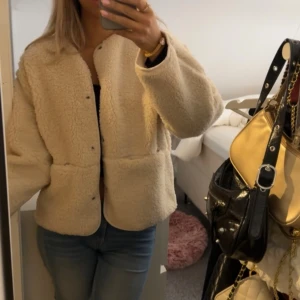 Beige teddyjacka med tryckknappar - Mysig beige teddyjacka med rund halsringning och stora fickor framtill. Jackan har tryckknappar och är lätt oversized i modellen, perfekt för lager-på-lager. Fodrad insida och lång ärm för extra komfort. Passar dig som gillar en chill och trendig look.