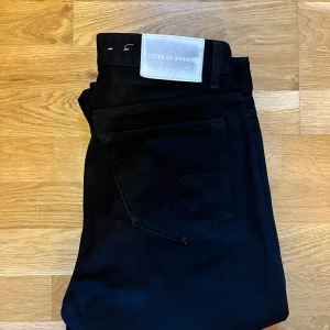 Svarta jeans från Tiger of Sweden - Svarta jeans från Tiger of Sweden med klassisk femficksdesign och slimfit design. Jeansen har en stilren look med svart tyg och diskret logotyp på bakfickan. Perfekta för dig som gillar grisch stilen.