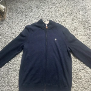 Morris kofta  - Mörkblå kofta från Morris med hel dragkedja och ribbad krage. Liten vit logga broderad på bröstet. Tillverkad i mjukt stickat material, perfekt för lager-på-lager. Klassisk och stilren design som funkar till det mesta.