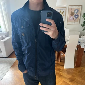 Blå vindjacka från Refrigwear - Säljer en stilren marinblå vindjacka från Refrigwear med dragkedja framtill och en bröstficka med knapp. Jackan har hög krage och klassisk passform, perfekt för vår och höst. Materialet är lätt och smidigt polyester.