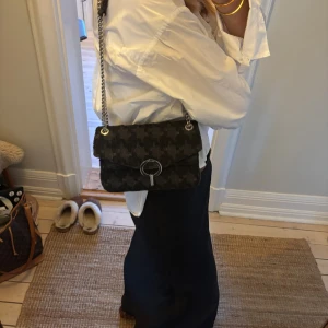 Svart axelväska Sandro med kedja - Snygg svart axelväska från Sandro med grovt rutigt mönster och silverfärgad kedja. Väskan har ett stort metallspänne framtill med Sandro-logga och är tillverkad i en mix av mocka och tyg. Köpt i USA på Sandro, jättefint skick!💕