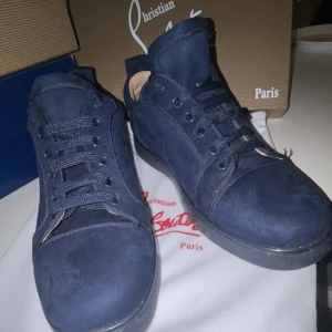 Mörkblå sneakers från Christian Louboutin - Snygga mörkblå sneakers från Christian Louboutin i mocka med klassisk röd sula. Skorna har rund tå, platt sula och snörning framtill. Insidan är ljusbeige och detaljerna är ton-i-ton. Perfekta för dig som vill sticka ut med exklusiva och trendiga skor.