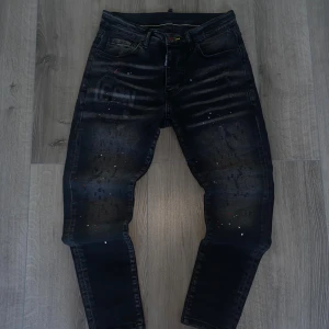 Svarta Dsquared2 ICON jeans med färgstänk - Snygga svarta jeans från Dsquared2 med ikonisk 'ICON'-logga på både fram- och baksida. Jeansen har skinny passform, slitningar och coola färgstänk i vitt och rött för en edgy vibe. Tillverkade i stretchig denim som sitter tajt och bekvämt.