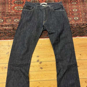 Mörkblå jeans från J.Lindeberg - Säljer ett par mörkblå jeans från J.Lindeberg med raka ben och klassisk femficksdesign. Jeansen har kontrastsömmar och tydliga bakfickor med märkets signaturbrodyr. Perfekta för dig som gillar stilrena och tidlösa jeans.