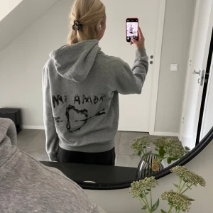 Grå hoodie mira paris - Grå hoodie med huva och snörning, samt känguruficka framtill. På ryggen finns ett svart tryck med texten 'Mi Amor' och ett hjärta genomborrat av en pil. Perfekt för dig som gillar en chill och avslappnad stil.