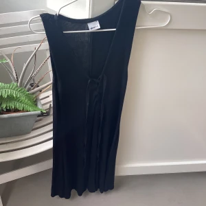 Svart långklänning med snörning Storlek S - Svart långklänning med snygg v-ringning och snörningsdetalj framtill. Klänningen är ärmlös och har en mjuk, följsam passform som ger en clean och stilren look. Perfekt för dig som gillar minimalistisk stil och vill ha något enkelt men ändå coolt.