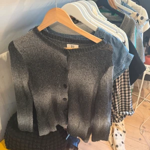 Grå kofta från Noa Noa med knappar - Snygg gråmelerad stickad kofta från Noa Noa i storlek M. Koftan har rund hals, långa ärmar och knäppning med svarta knappar framtill. Mjuk och skön känsla, perfekt för lager-på-lager. Passar dig som gillar klassisk stil med en twist.