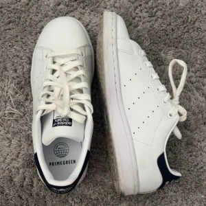 Stan Smith  - Klassiska Adidas Stan Smith sneakers i vitt med mörkblå detalj på hälen. Skorna har snörning, rund tå och är tillverkade i syntetmaterial. Perfekta för dig som gillar en clean och tidlös stil.