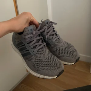 Grå Adidas Ultraboost sneakers - Säljer ett par grå Adidas Ultraboost sneakers med stickad ovandel och klassiska tre ränder på sidorna. Skorna har snörning, vit Boost-sula och detaljer i mörkgrått. Perfekta för dig som gillar stilrena och bekväma sneakers till vardags eller träning. 