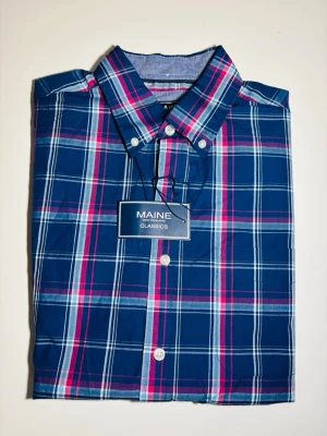 Blårutig kortärmad skjorta Maine - Kortärmad skjorta från Maine med blå, rosa och vita rutor. Klassisk button-down krage och vita knappar framtill. Tillverkad i bomull för en skön känsla. Perfekt för dig som gillar färg och mönster i din stil.