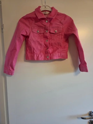 Rosa jeansjacka från H&M, stl 128 - Snygg rosa jeansjacka från H&M i klassisk modell med krage, silverfärgade knappar och två bröstfickor. Jackan är croppad och har långa ärmar. Perfekt för dig som vill sticka ut med färg och gillar streetstyle.